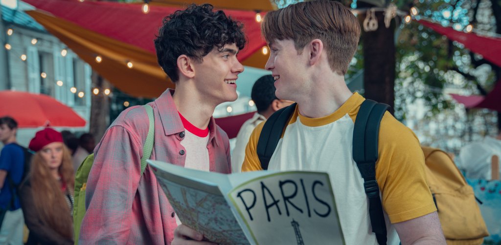 Kit Connor e Joe Locke como Nick e Charlie na 2ª temporada de Heartstopper; eles estão sorrindo enquanto olham um para o outro com um panfleto de Paris nas mãos