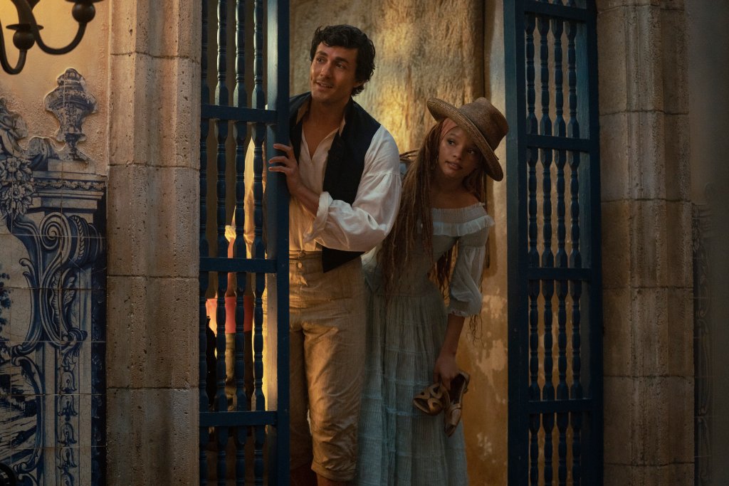 Jonah Hauer-King como príncipe Eric e Halle Bailey como Ariel no live-action de A Pequena Sereia; eles estão sorrindo enquanto se inclinam para olhar algo em uma passagem