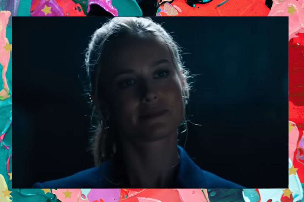 Brie Larson em cena de Velozes & Furiosos 10 sorrindo levemente com o cabelo preso; a margem é uma textura nas cores lilás, rosa, vermelho, roxo, verde e preto; estrelas amarelas decoram a imagem