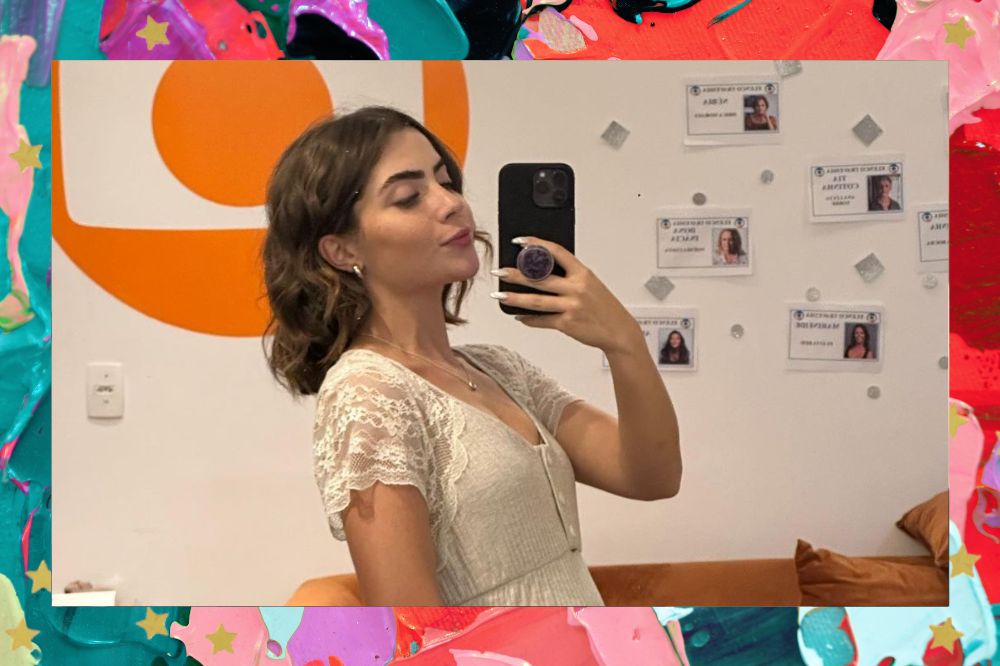 Jade Picon tirando uma foto no espelho com uma parede branca com fotos e o logo da Globo em laranja ao fundo; ela usa um vestido branco com detalhes de renda; a margem é uma textura nas cores lilás, verde, roxo, vermelho e rosa com estrelas amarelas como decoração