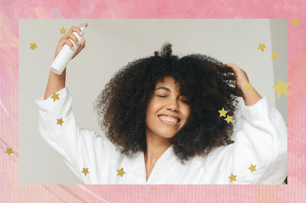 Foto de garota de cabelo crespo com olhos fechados, sorrindo, usando roupão branco enquanto passa spray de cabelo com uma das mãos. Montagem tem fundo rosa com estrelinhas douradas