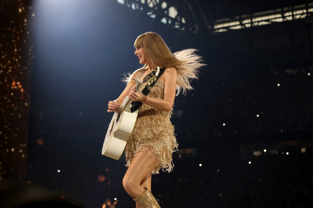 Taylor Swift na The Eras Tour; ela está tocando violão no palco enquanto sorri e roda dançando com um vestido dourado de franjas