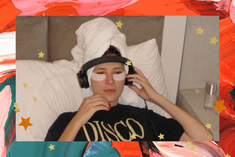 Montegem com fundo em tons laranja, rosa e verde. Mulher no centro deitada em uma cama com uma toalha na cabeça, um fone de ouvido e mascara de hidratação embaixo dos olhos.