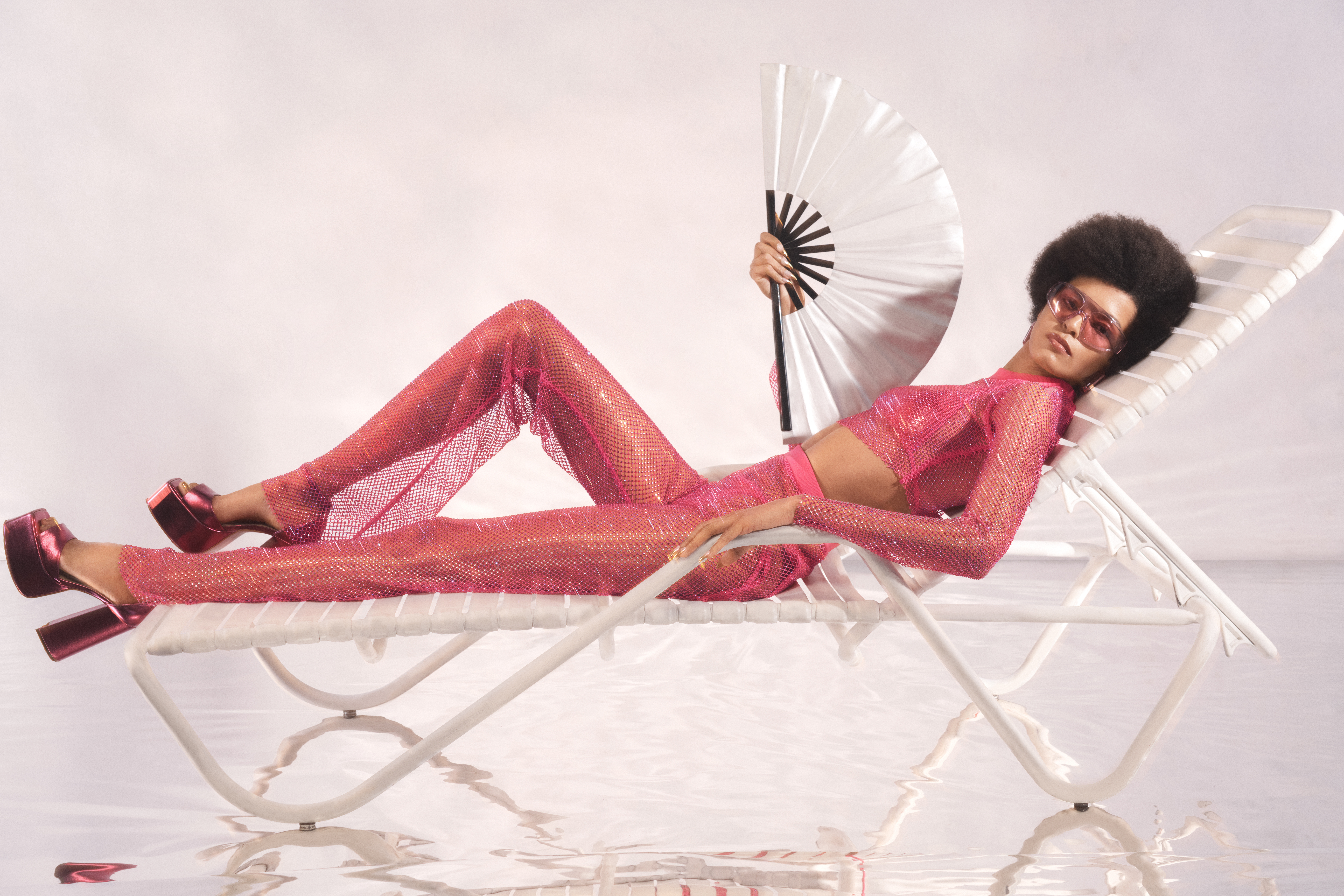 Laiza de Moura está deitada em uma cadeira de piscina branca e segura um leque branco. Ela usa calça, cropped de manga comprida, óculos de sol e salto plataforma rosas. Seu olhar está direcionado para a câmera.