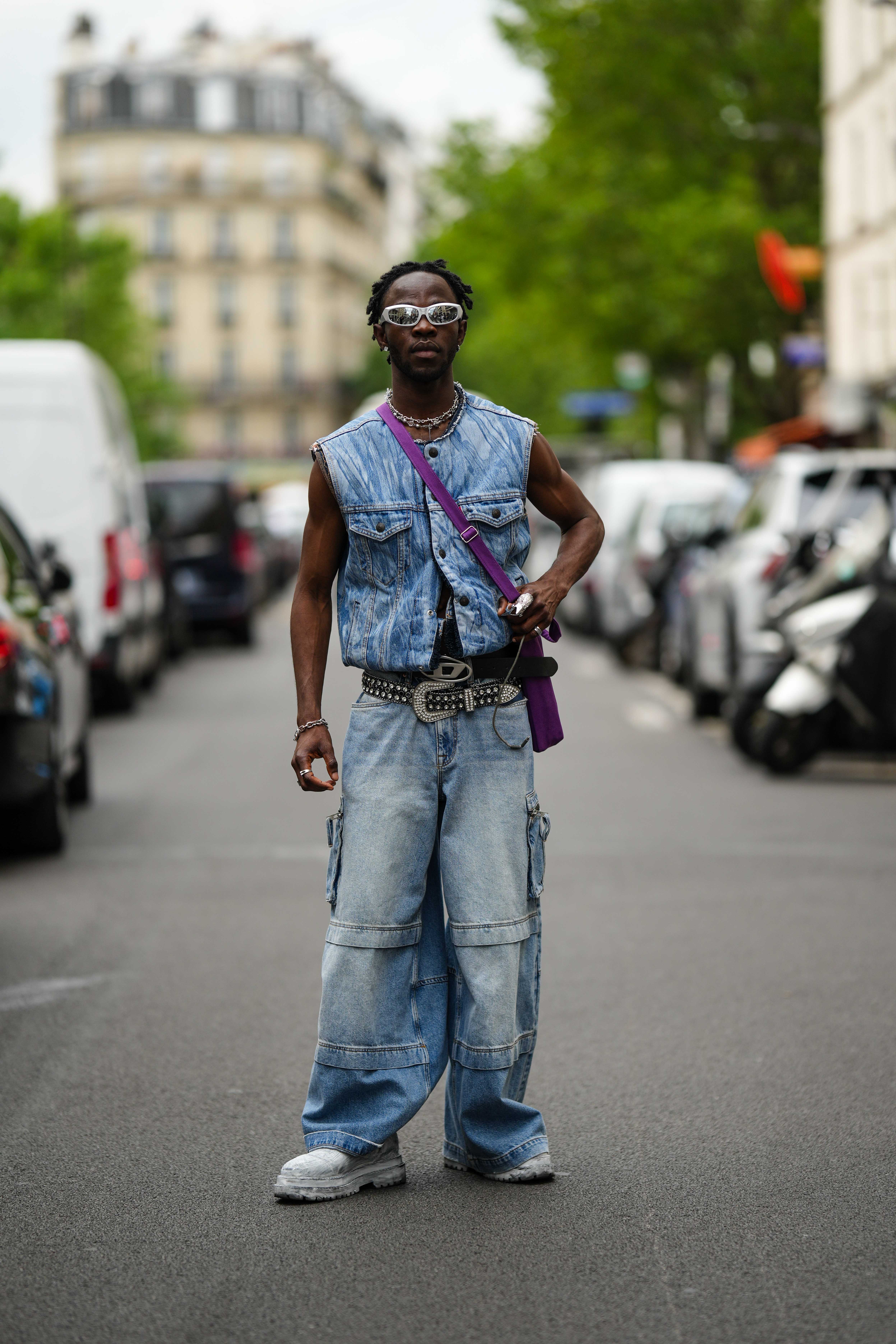 Look no street style da semana de moda masculina de primavera-verão 2024 em Paris. Homem usando colete e calça cargo jeans, tênis branco, mix de cintos, bolsa roxa e óculos metalizados