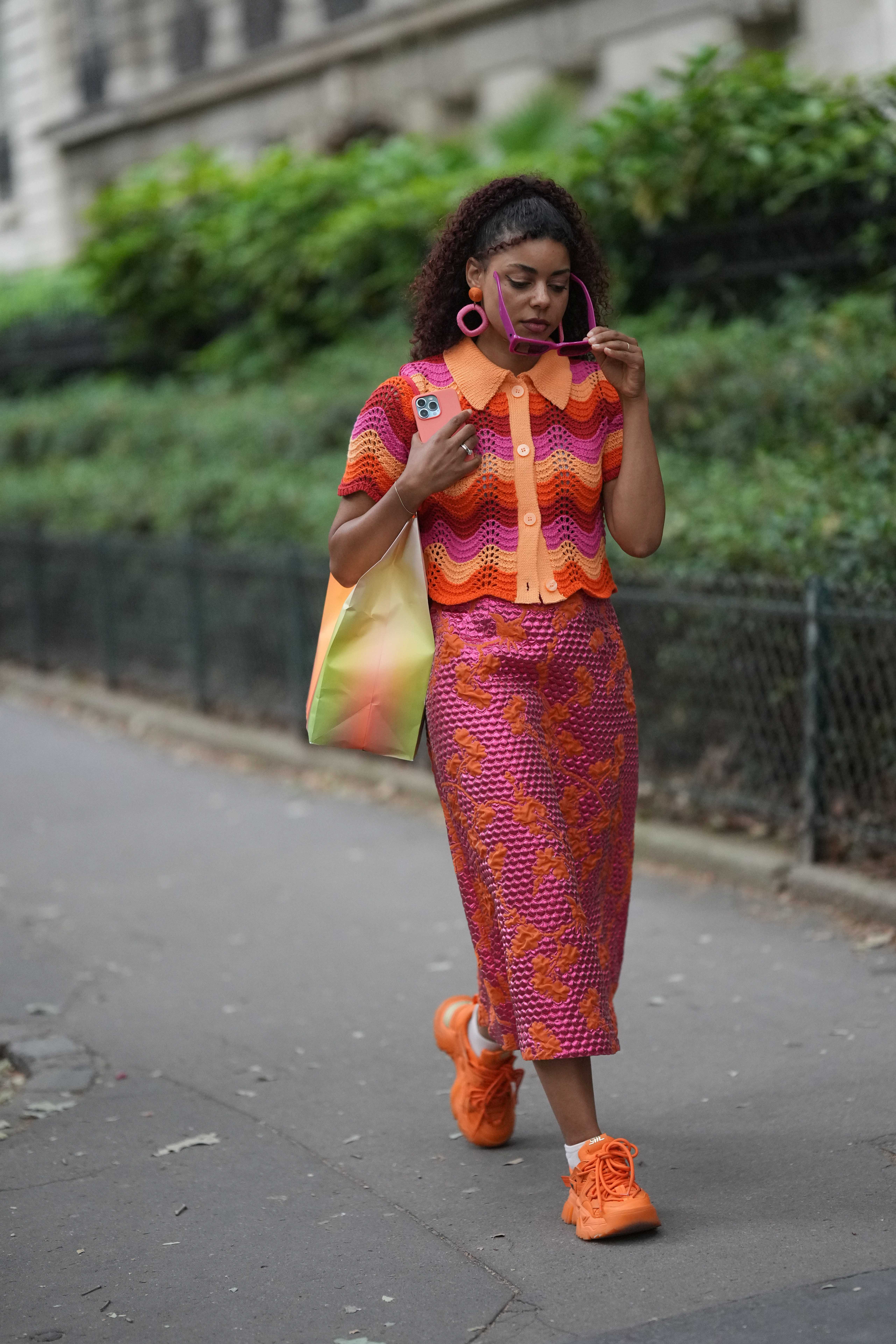 Look no street style da semana de moda masculina de primavera-verão 2024 em Paris. Mulher usando look todo rosa e laranja com blusa de tricô, saia midi e tênis esportivo