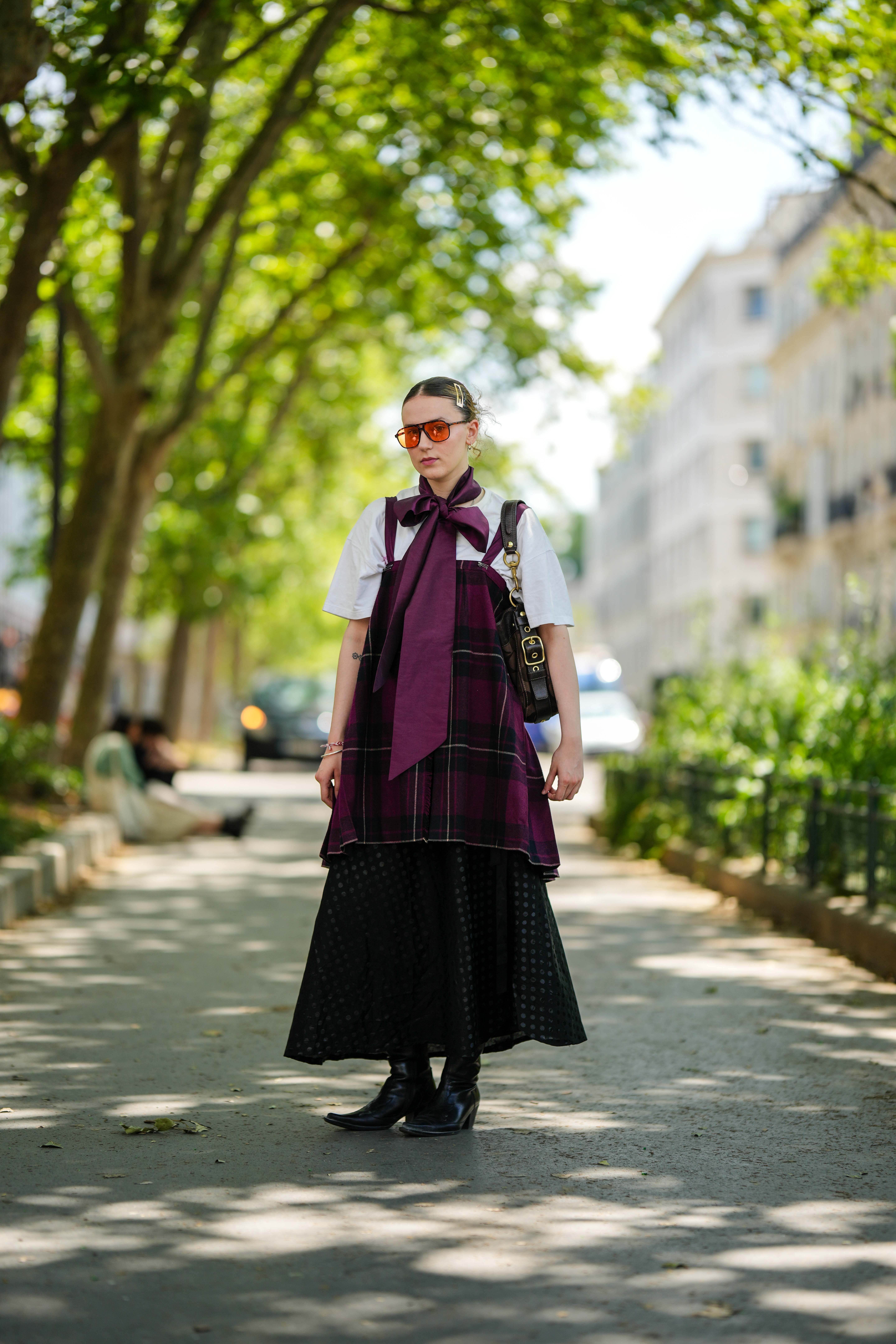 Look no street style da semana de moda masculina de primavera-verão 2024 em Paris. Mulher usando camiseta branca sobreposta a peça xadrez e saia com bota western preta e óculos de lentes amareladas