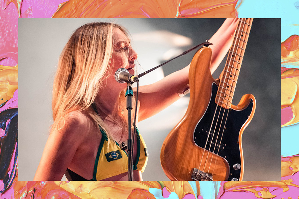 Este Haim em show no MITA Festival usando biquíni do Brasil e segurando o baixo enquanto canta. Montagem em fundo laranja, amarelo, rosa e azul