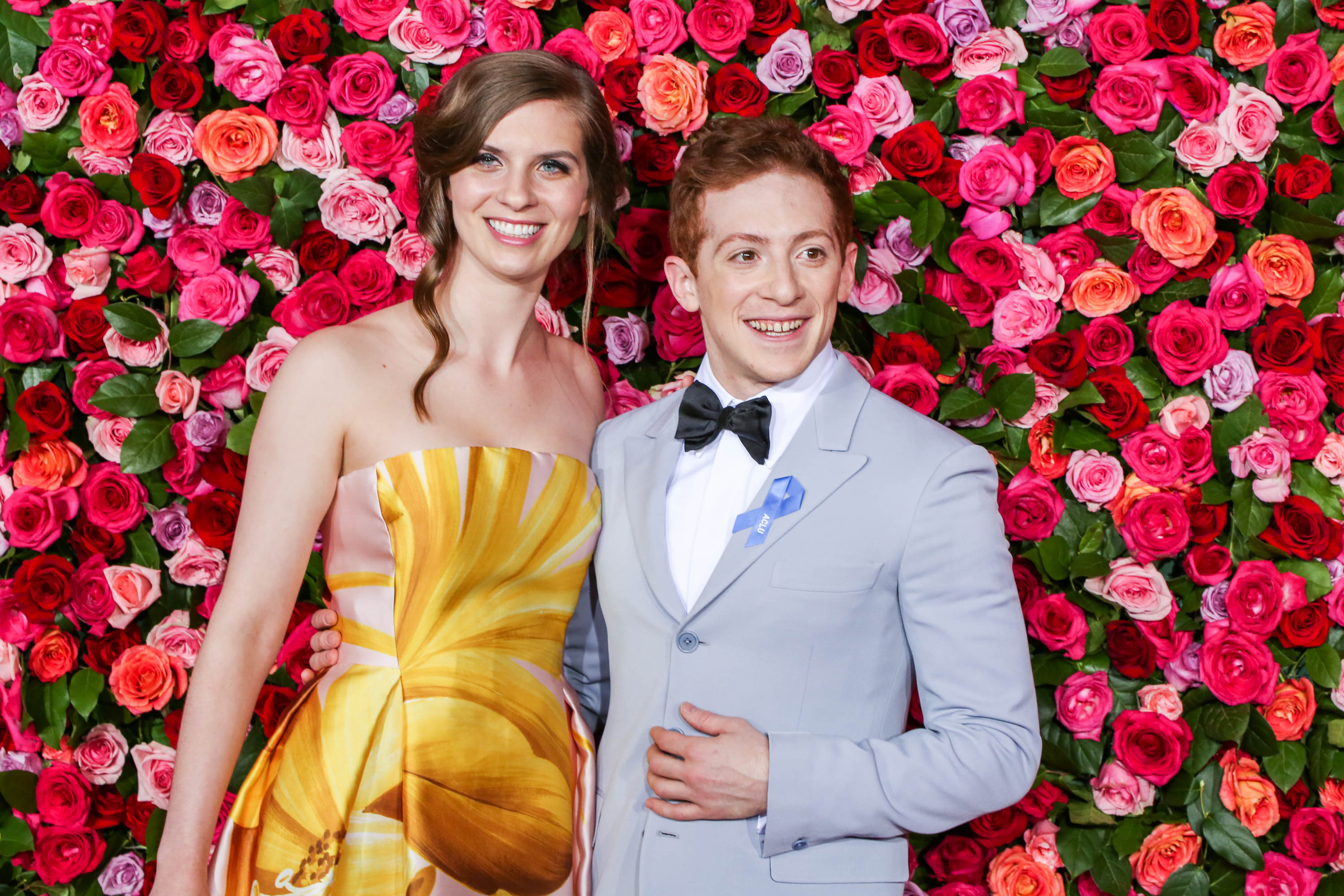 Ethan Slater e Lilly Jay sorrindo enquanto posam para foto com uma parede de flores coloridas ao fundo