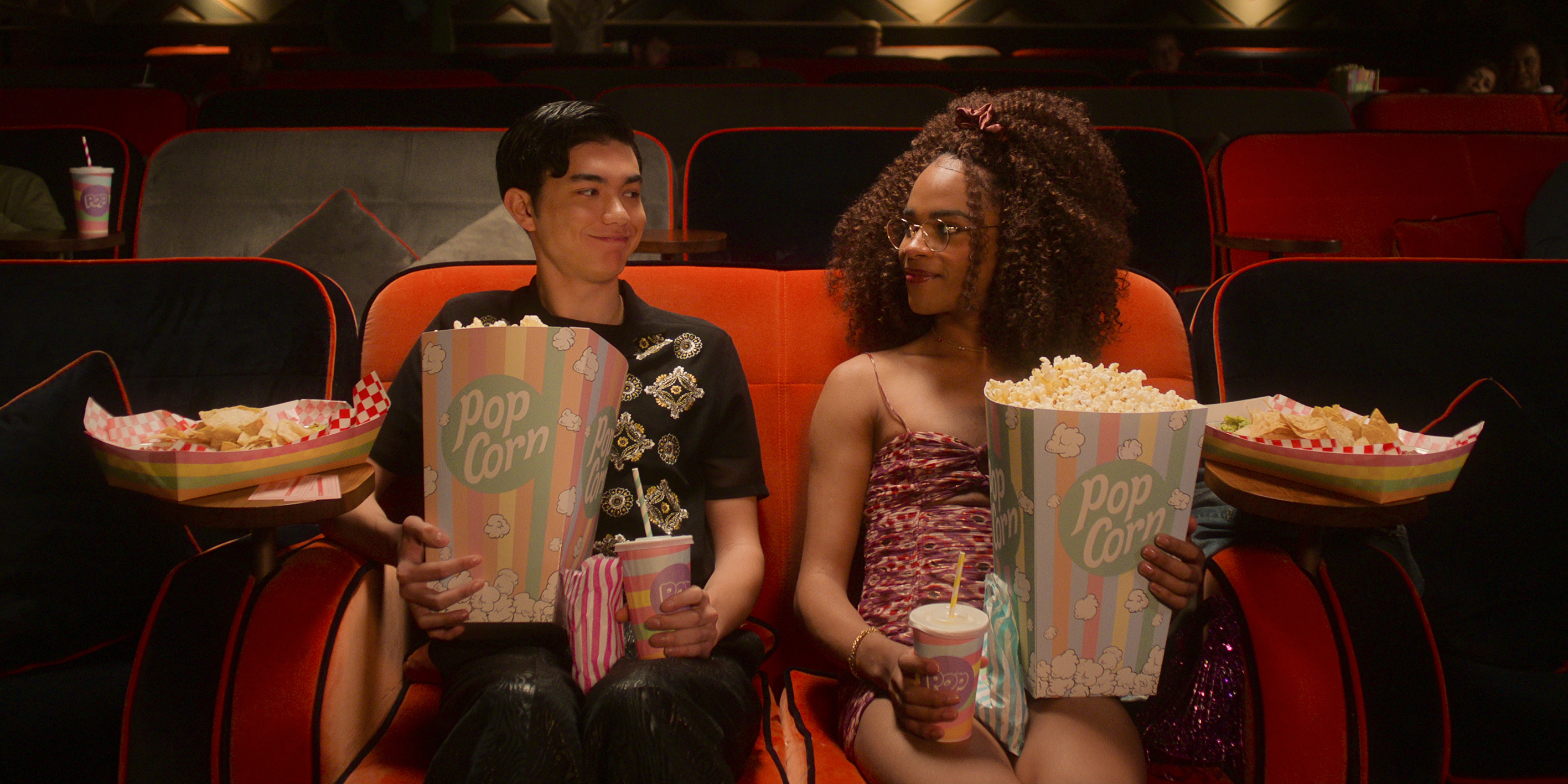 Will Gao e Yasmin Finney em cena de Heartstopper; eles estão no cinema se olhando enquanto seguram grandes pacotes de pipoca e bebidas; eles sorriem levemente um para o outro