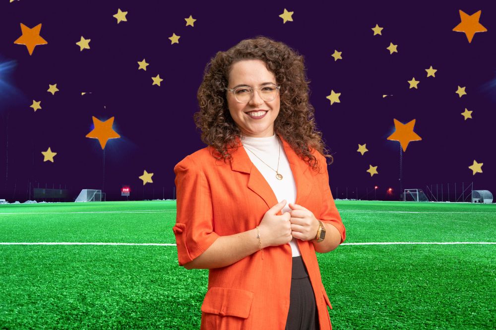 Natália Lara, uma mulher branca de cabelos castanhos cacheados, está vestindo um blazer laranja. O fundo da foto é um campo de futebol com o céu azul e estrelas