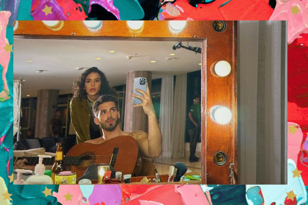 Imagem de Bruna Marquezine e João Guilherme posando para foto no espelho em camarim; ela está em pé atrás dele, que está sentado e segurando um violão; a margem é uma textura nas cores lilás, azul, roxo, verde, rosa e vermelho; estrelas amarelas decoram a imagem