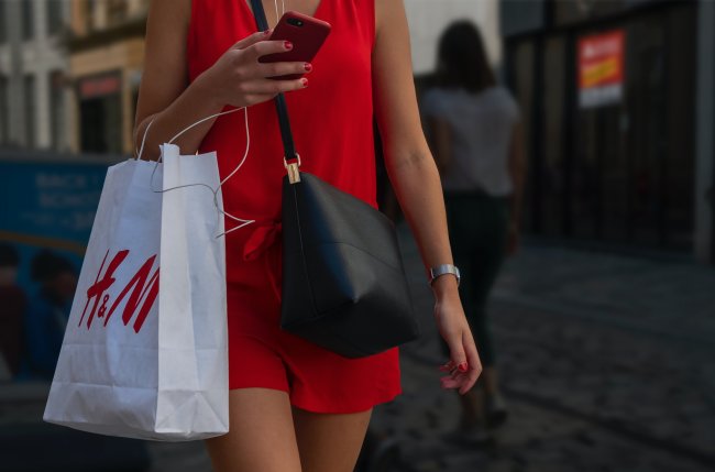 Mulher com vestido vermelho e bolsa preta carregando uma sacola de compras da H&M e segurando o celular. Não dá para ver a cara.