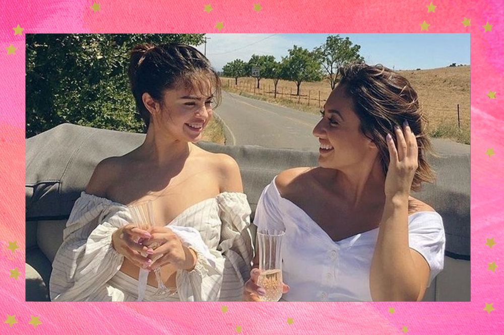 Selena Gomez e Francia Raísa olhando para uma outra e sorrindo; ambas usam roupas brancas e estão segurando copos em um ambiente externo e com árvores ao fundo durante o dia; a margem é uma textura em tons de rosa e branco com estrelas amarelas como decoração