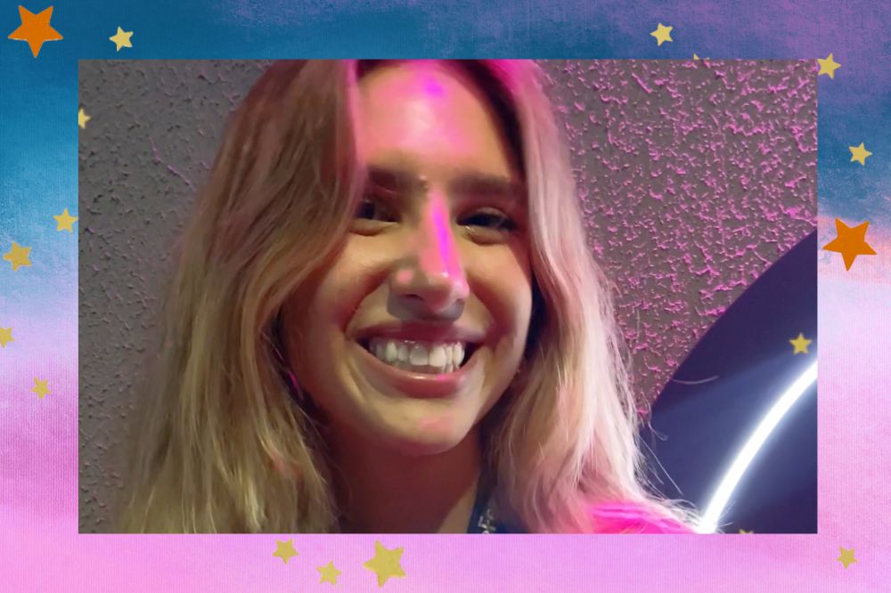 Marcela Montellato sorrindo com iluminação rosa sendo refletida em seu rosto; a margem é uma textura nos tons azul, rosa e branco com estrelas amarelas e laranjas decorando a imagem