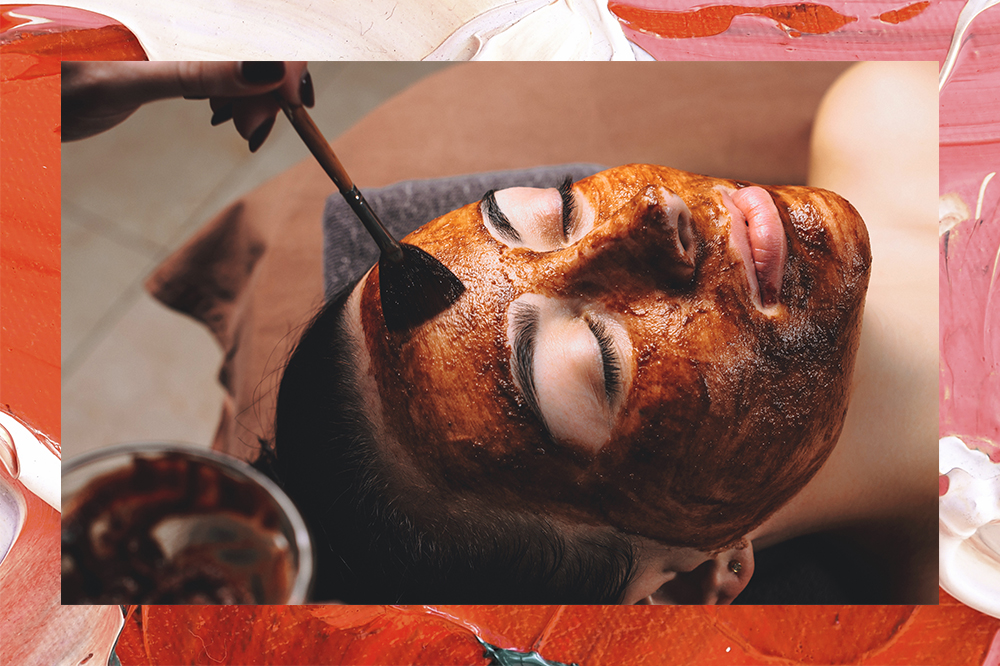 Mulher deitada com os olhos fechados enquanto uma mão passa uma máscara facial de chocolate em seu rosto