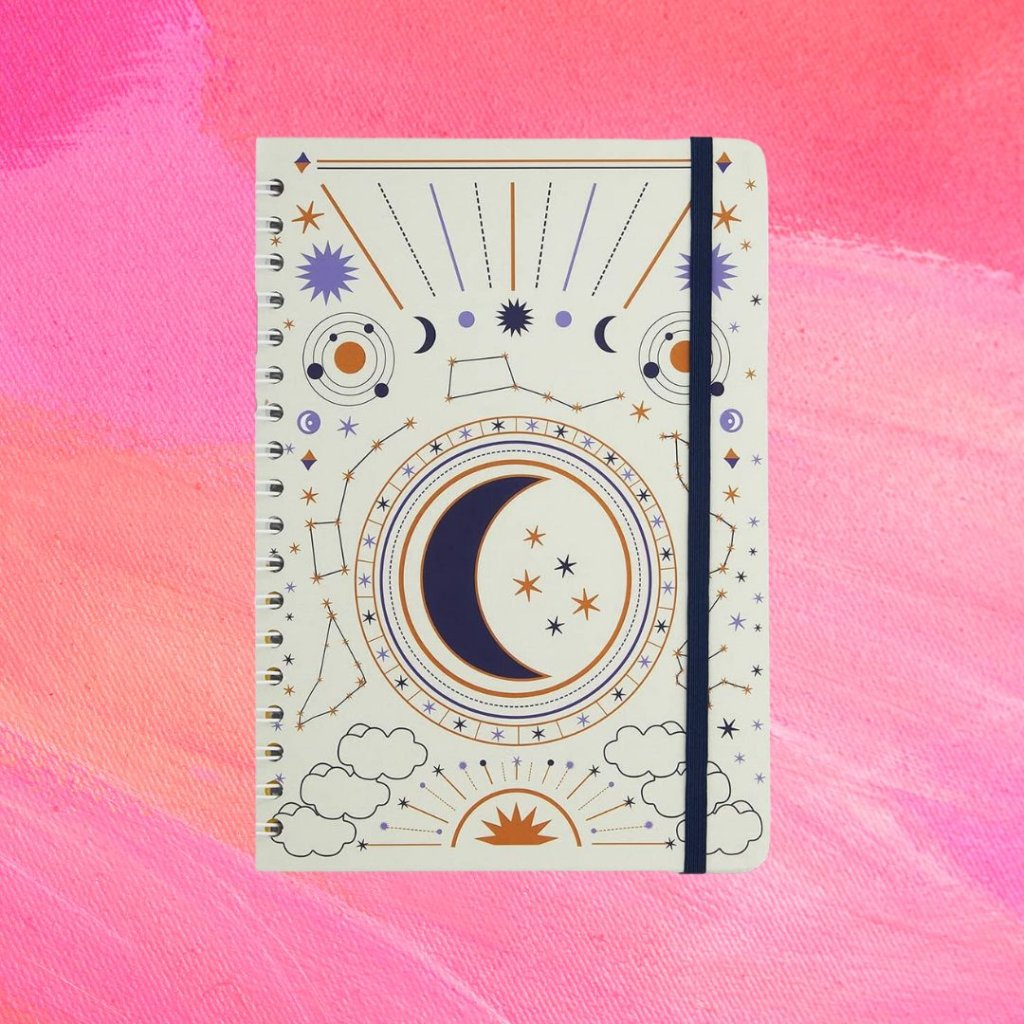 Caderno branco com estampa de sol, lua e estrelas em azul, dourado e roxo e uma tira azul escura; o fundo é uma textura em tons de rosa e branco