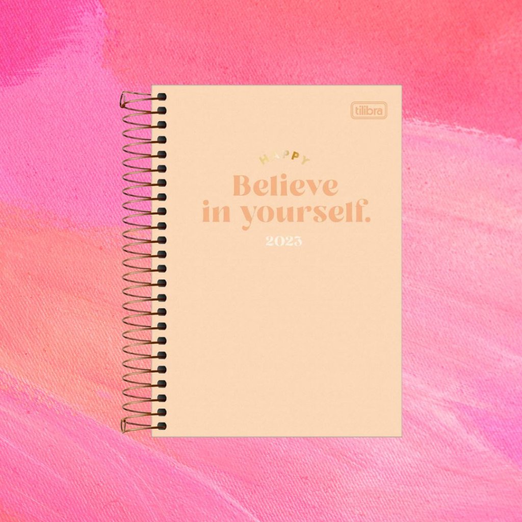Planner rosa com texto "Happy/Believe In Yourself/2023" em dourado, rosa e branco; o fundo é uma textura em tons de rosa e branco
