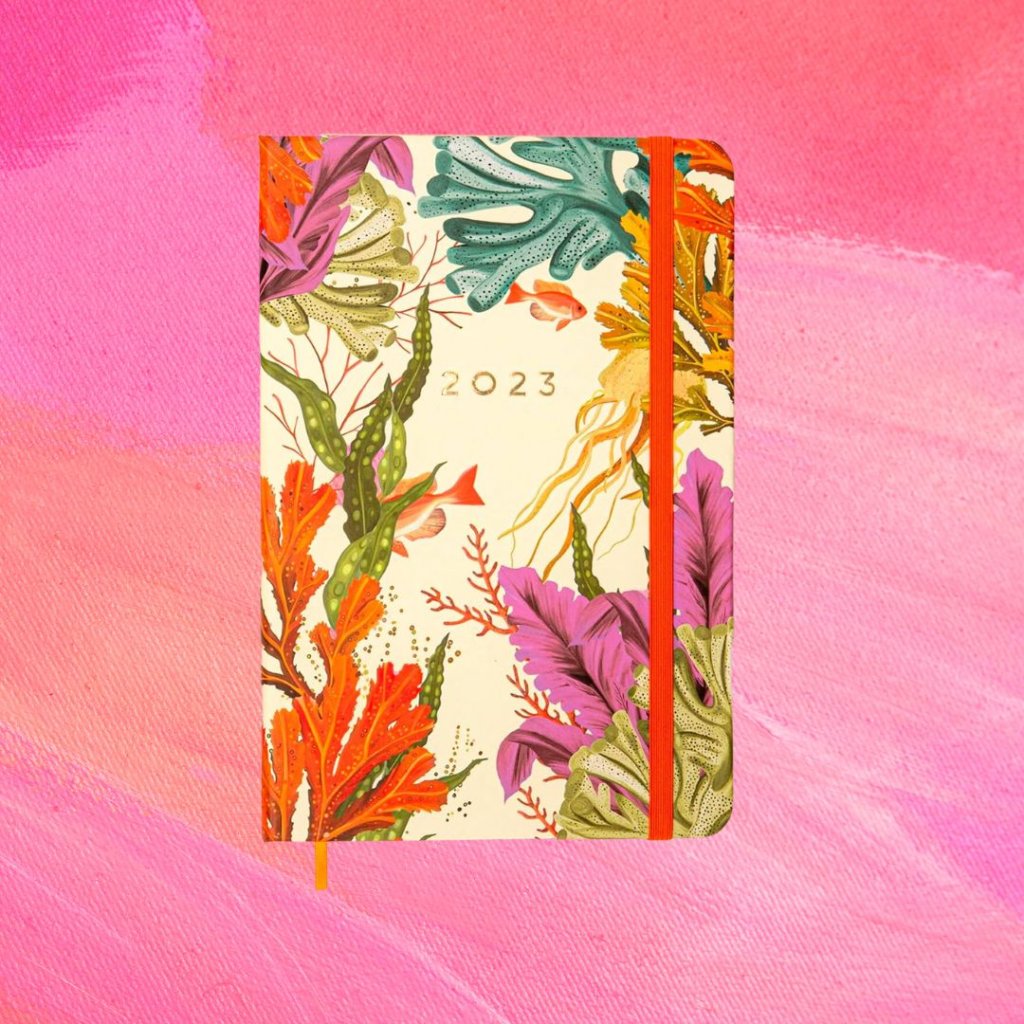 Planner florido em diferentes cores com o texto 2023 em dourado na parte central da capa e uma tira laranja; o fundo é uma textura em tons de rosa e branco