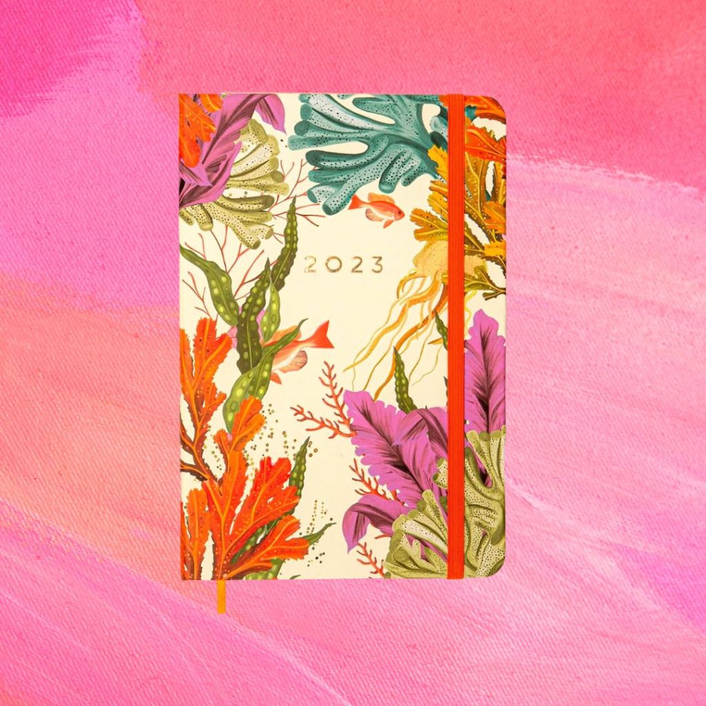 planner (8) Planner florido em diferentes cores com o texto 2023 em dourado na parte central da capa e uma tira laranja; o fundo é uma textura em tons de rosa e branco