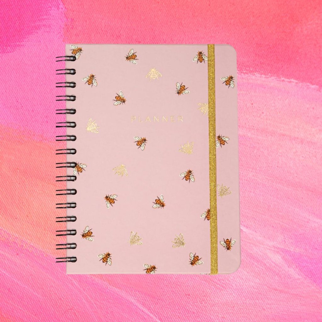 Planner rosa com detalhes de abelhas na estampa e uma tira dourada; o fundo é uma textura em tons de rosa e branco