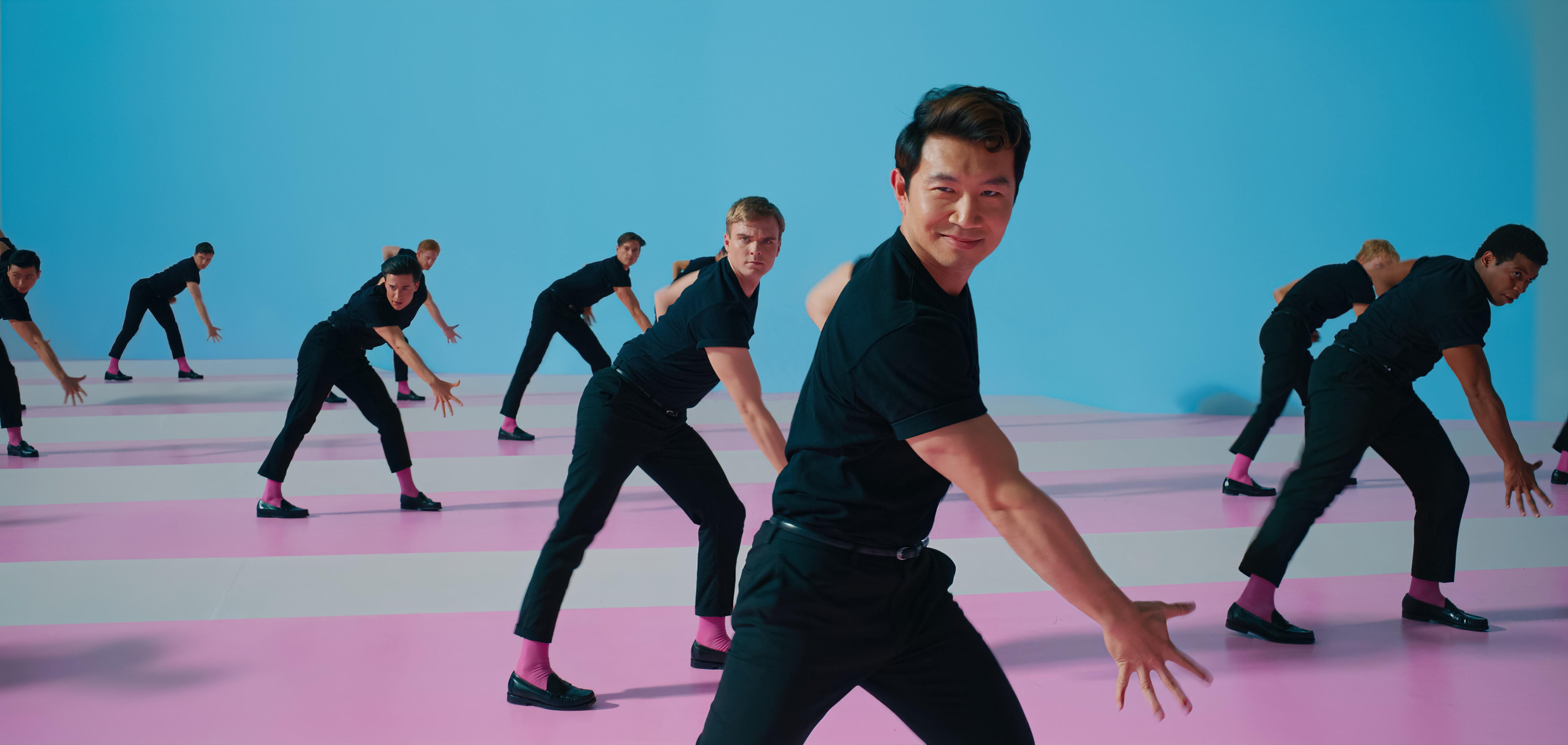 Simu Liu dançando em cena de Barbie; ele está usando preto e fazendo a coreografia com outros Kens ao fundo também dançando