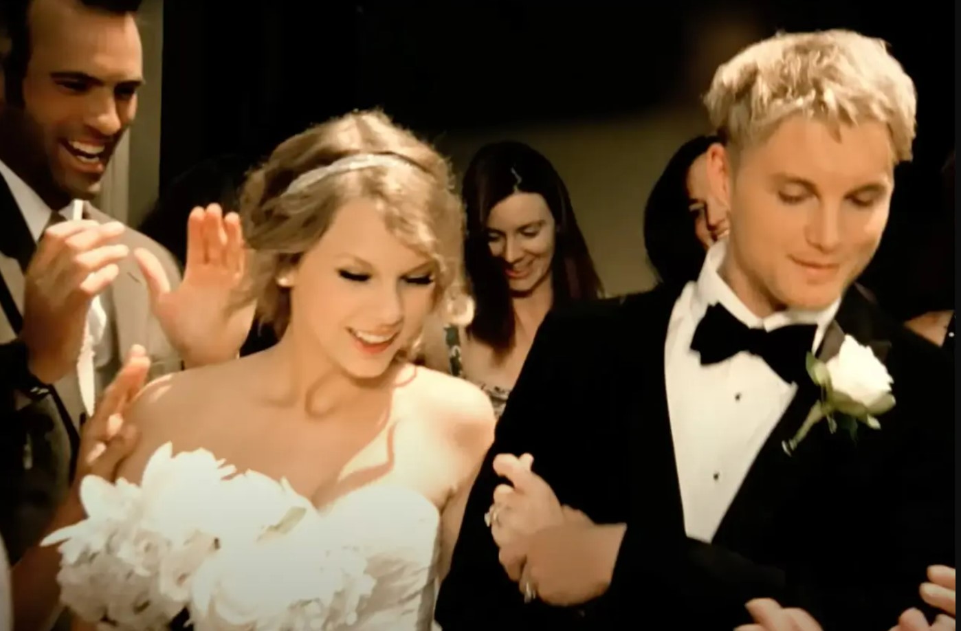 Taylor Swift vestida de noiva com um buquê de flores brancas na mão e um vestido tomara que caia branco. Um homem louro - como um noivo - ao lado com um smooking preto. A cena é do clipe de Mine.