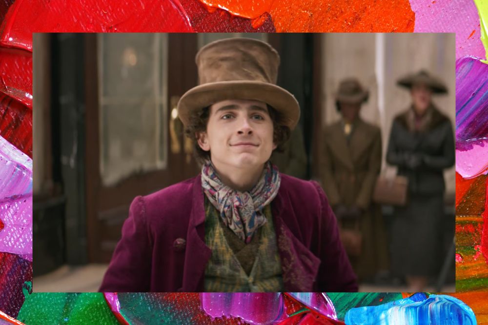 Timothée Chalamet sorrindo levemente em cena do trailer de WONKA; ele está de chapéu e caracterizado como o personagem; a margem é uma textura de tintas nas cores vermelho, rosa, amarelo, roxo, azul, verde e branco