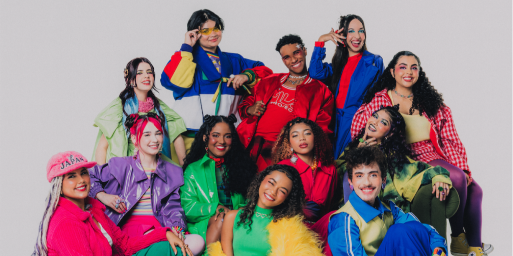 Grupo de 11 jovens sorrindo, com roupas coloridas e estilosas, posando em um fundo branco. Eles exibem maquiagens vibrantes e acessórios marcantes, transmitindo alegria e diversidade.