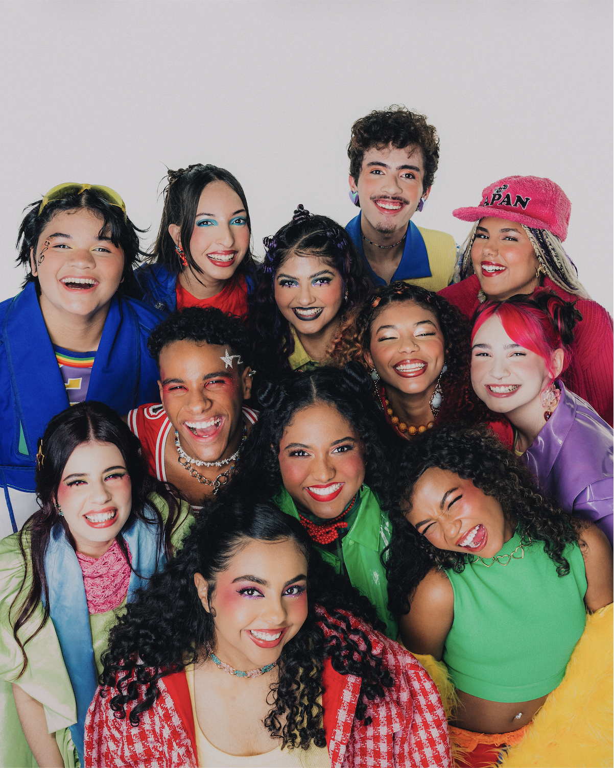 Grupo de doze jovens sorridentes, com maquiagens coloridas e roupas vibrantes, posando juntos em fundo branco. Eles exibem estilos diversos, com cabelos cacheados, lisos, coloridos e penteados variados, transmitindo alegria e diversidade