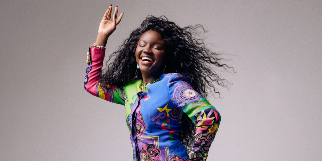 Mulher negra de cabelos longos e cacheados, sorrindo com os olhos fechados e a boca aberta, vestindo um blazer colorido com estampas psicodélicas, levantando a mão direita com a palma aberta, em fundo cinza claro