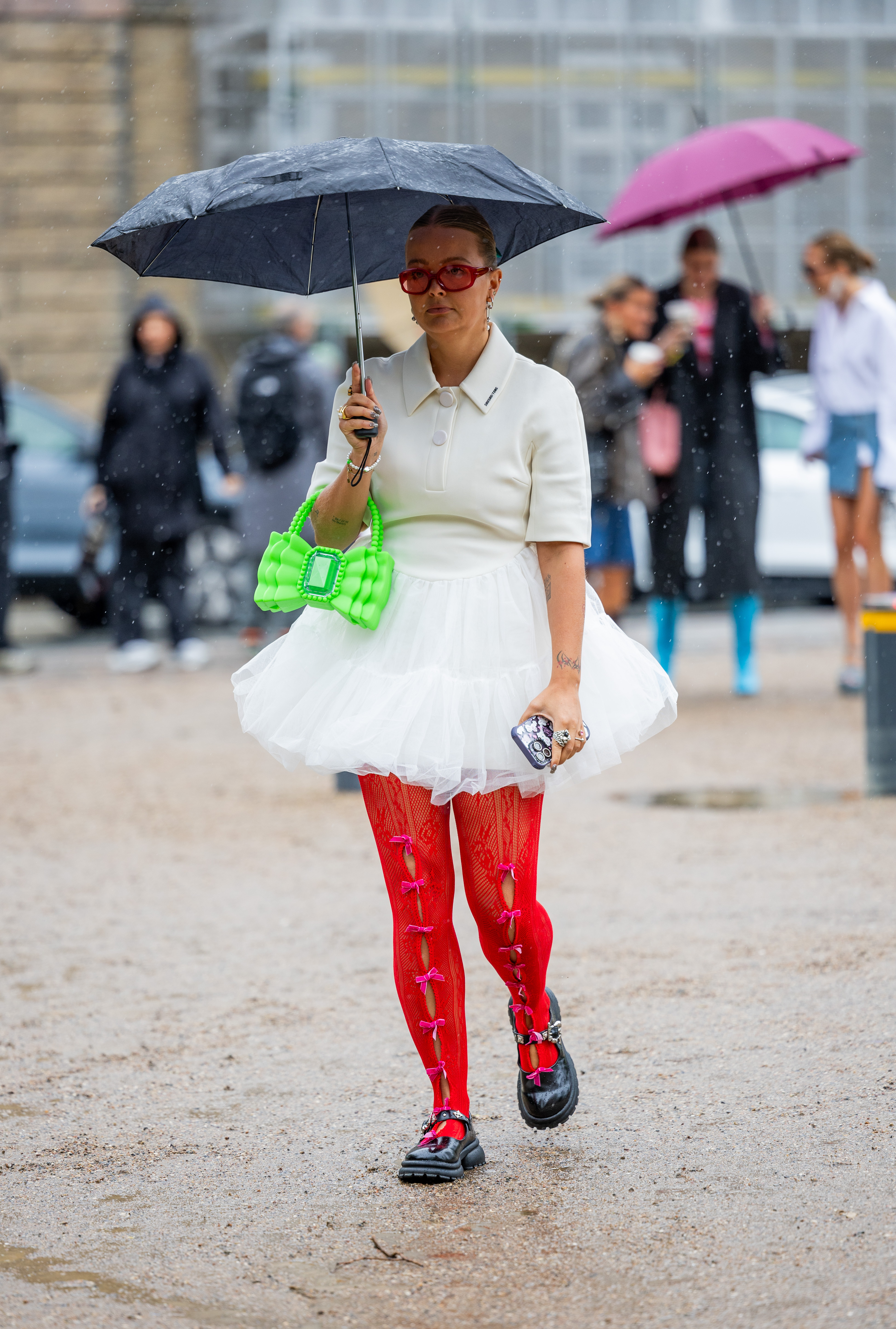 Look da semana de moda de Copenhagen na temporada de primavera-verão 2024 com blusa e saia off white, bolsa verde neon, meia-calça vermelha com laços e sapato preto