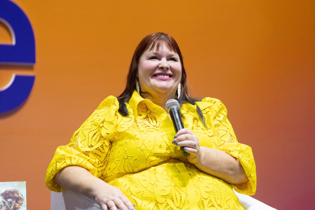 Cassandra Clare sentada e sorrindo com vestido amarelo enquanto segura um microfone