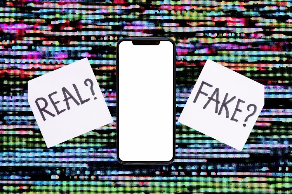 Fundo colorido. No meio, um smartphone com a tela branco e de cada lado um post-it branco com os escritos "Real?" ou "Fake?"