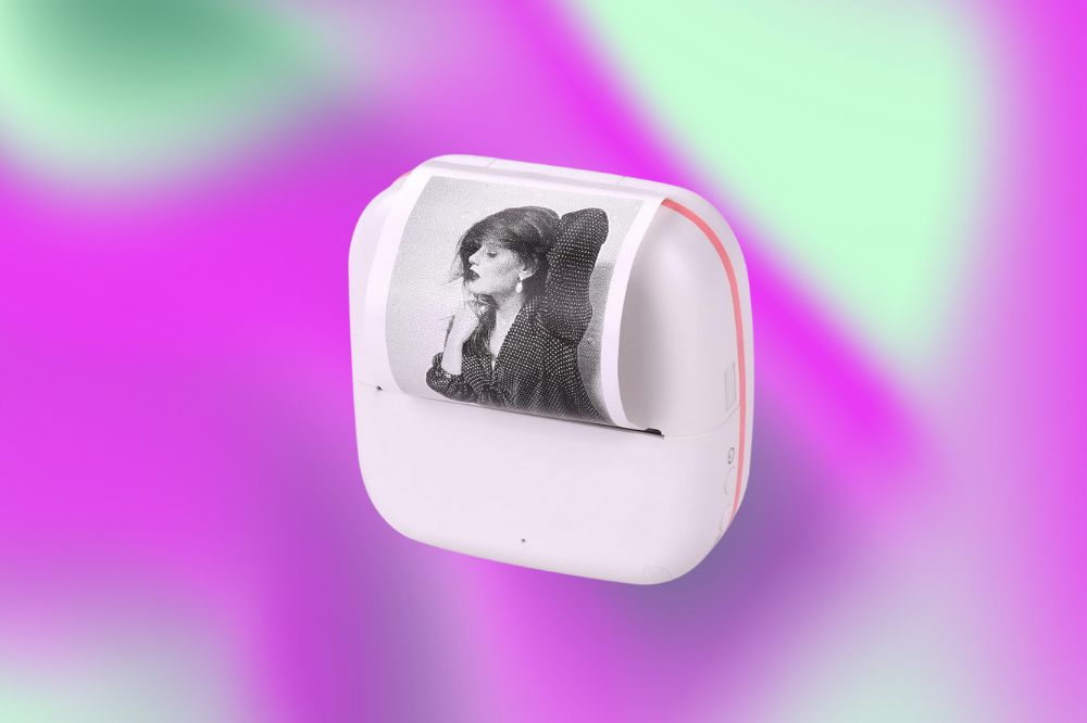 Uma impressora de fotos pequena, branca com detalhes rosa com um formato arredondado. Dela está sendo revelada uma foto de uma mulher em preto e branco. O fundo da imagem é rosa, roxo e verde claro.