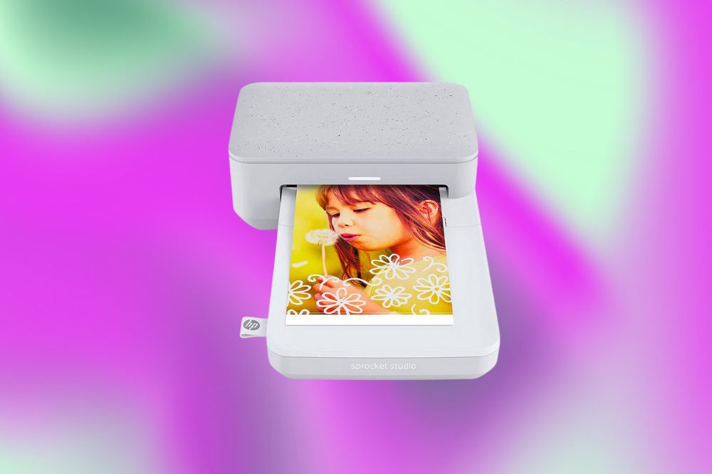 Uma impressora de fotos pequena branca com um formato arredondado. Dela está sendo revelada uma foto de uma garota com uma flor. O fundo da imagem é rosa, roxo e verde claro.