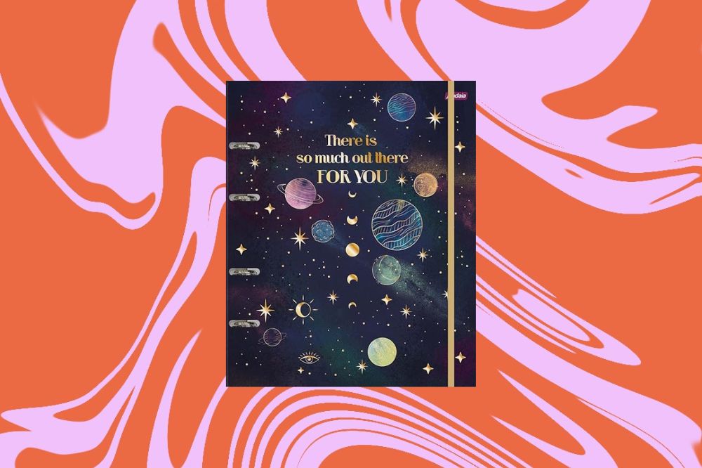 Caderno com estampa de galáxia, com planetas desenhados ao meio, em um fundo laranja e rosa