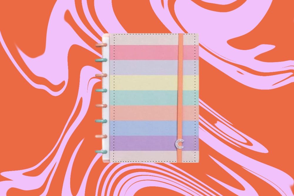 Caderno com estampa nas cores do arco-íris, em tom pastél, em um fundo laranja e rosa