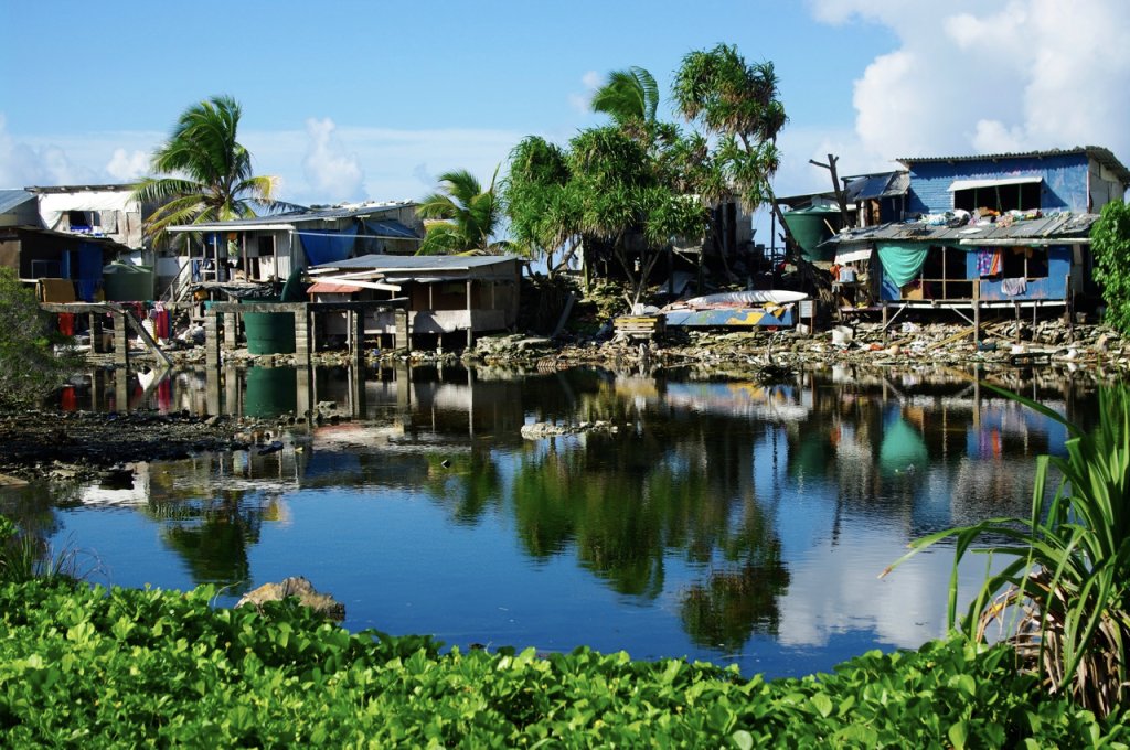 Na imagem, aparece casas construídas ao redor de um rio, lixos espalhados e coqueiros na parte de trás