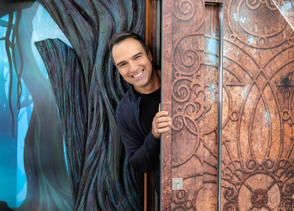 Tadeu Schmidt na casa do BBB24; ele está sorrindo enquanto espia a casa pela porta de entrada da área externa