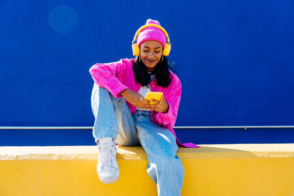 Garota negra de cabelos crespos e pele clara escuta música pelo celular com fones de ouvindo sentada em um muro amarelo e sorrindo.