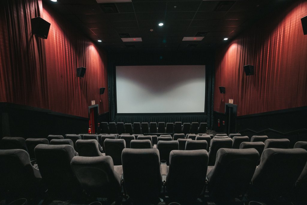 Sala de cinema com paredes vermelhas, cadeiras pretas e a tela branca ao fundo