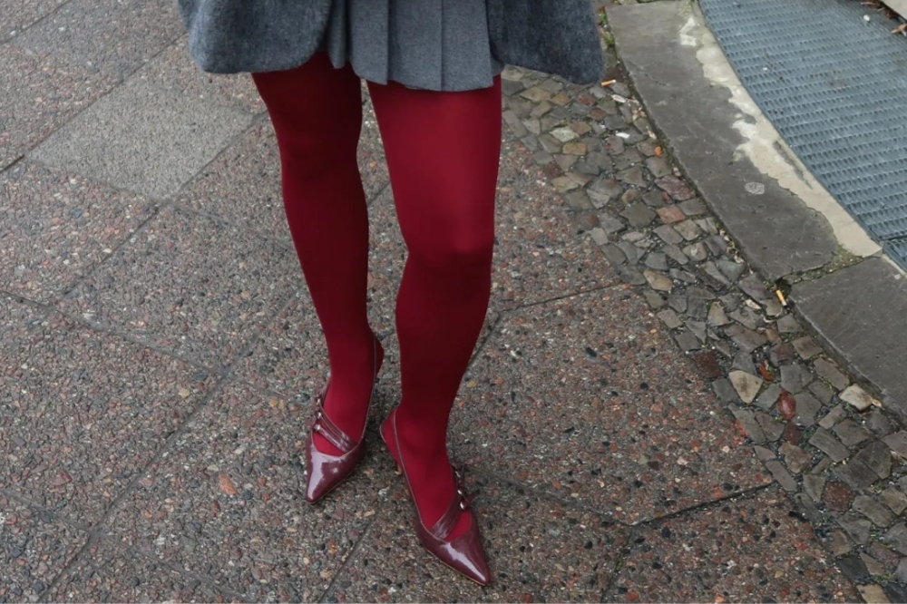 Perna de garota usando meia-calça vermelha bordô, sapato kitten heel e saia plissada cinza