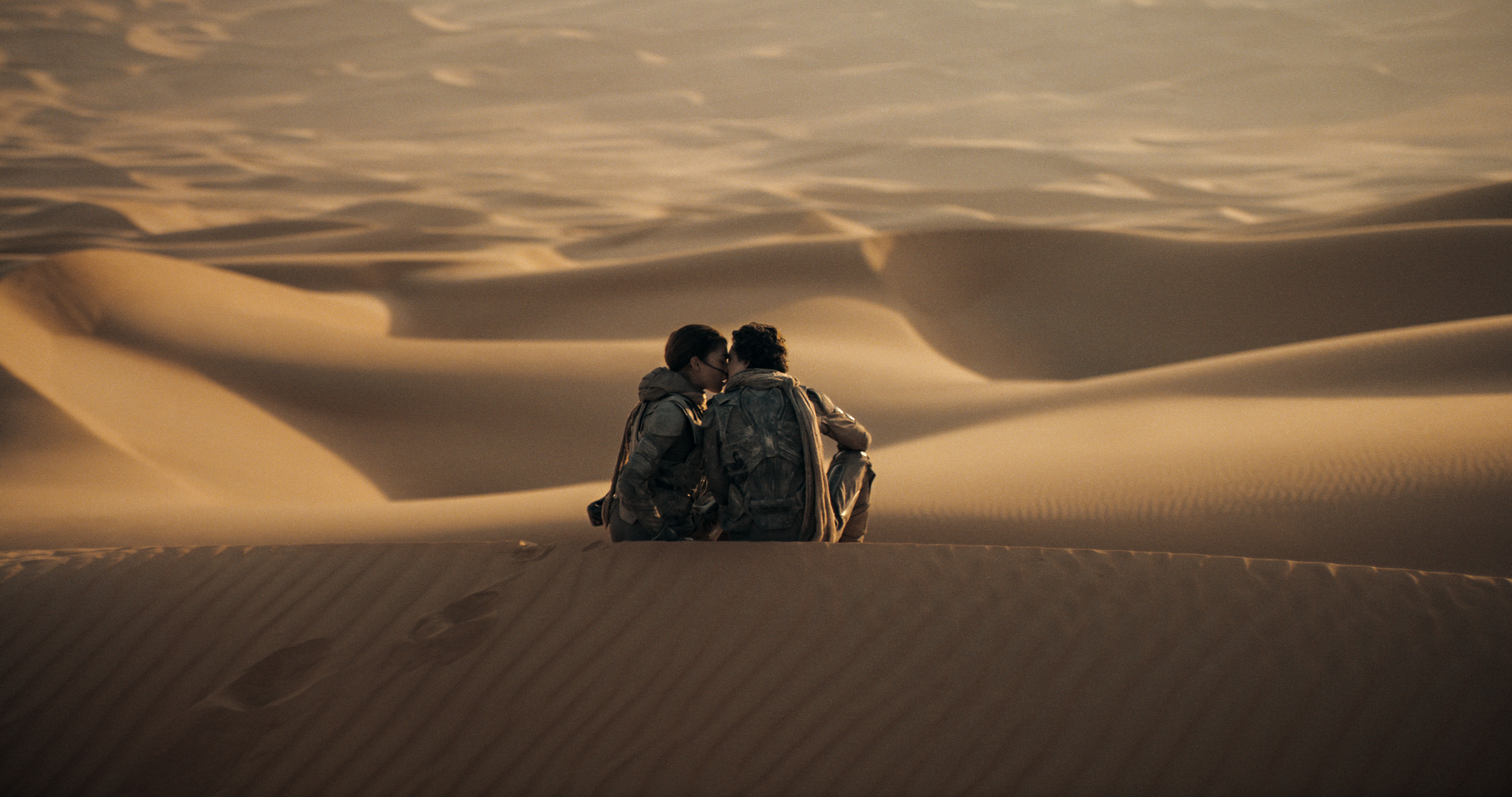 Zendaya e Timotheé Chalamet como Chani e Paul em cena de Duna Parte 2; eles estão no deserto sentados nas dunas de areia enquanto se beijam