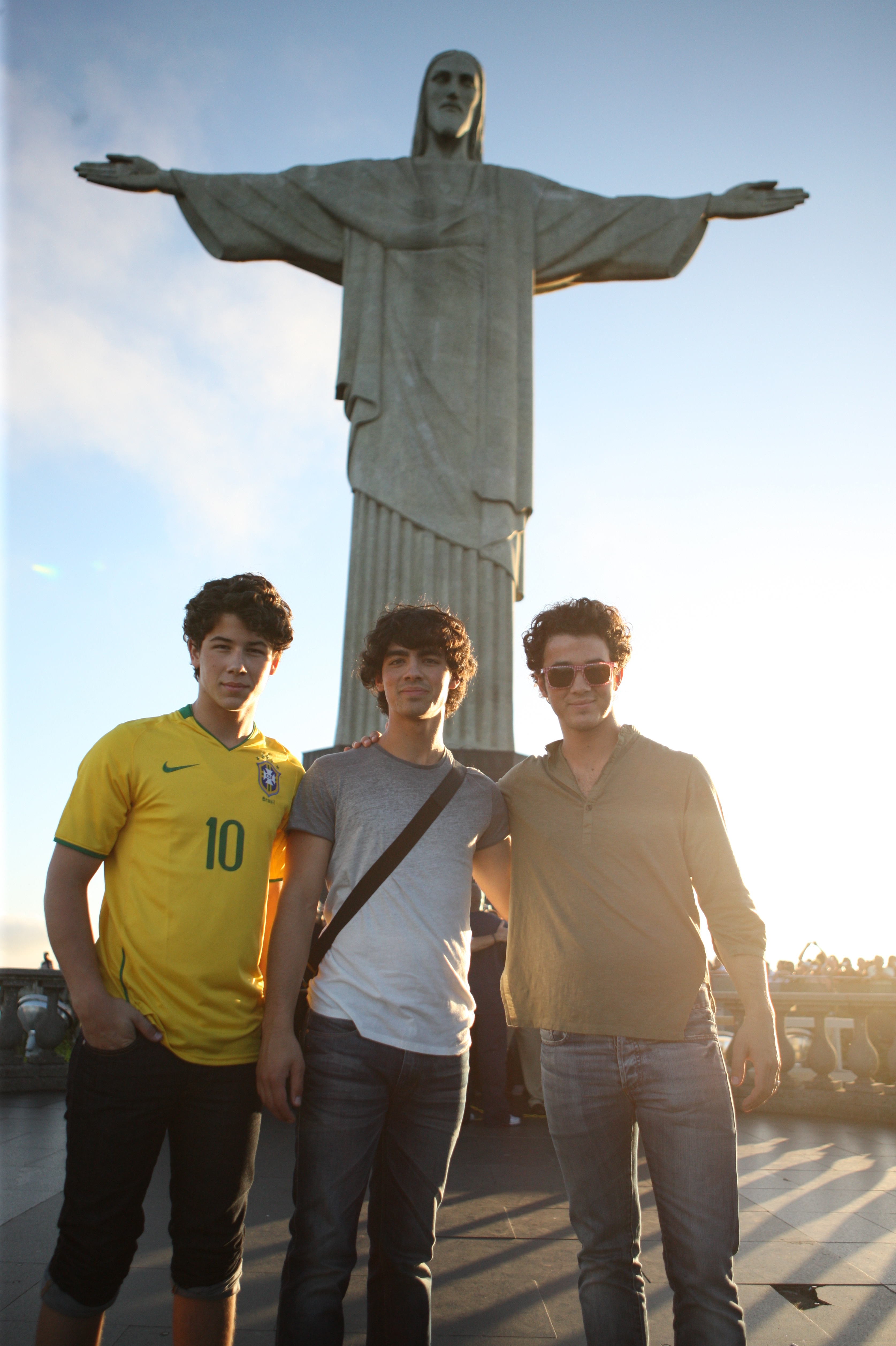 Nick, Joe e Kevin Jonas, dos Jonas Brothers, visitando o Cristo Redentor no Rio de Janeiro; eles estão sorrindo levemente e posando para foto na frente da estátua durante o dia