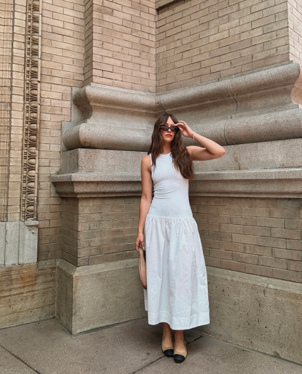 Garota usando vestido midi branco com sapatilha, óculos de sol e segurando bolsa bege na mão