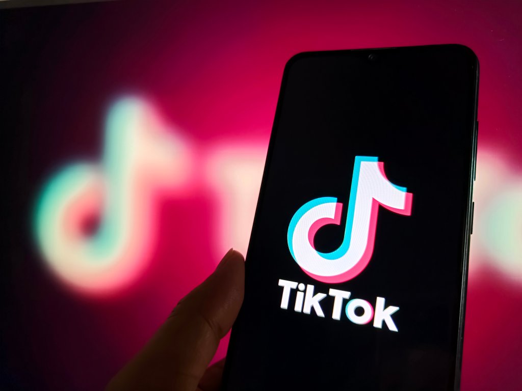 Imagem de uma mão segurando um celular com o logo do TikTok aberto e, ao fundo, é possível ver o logo do app borrado.