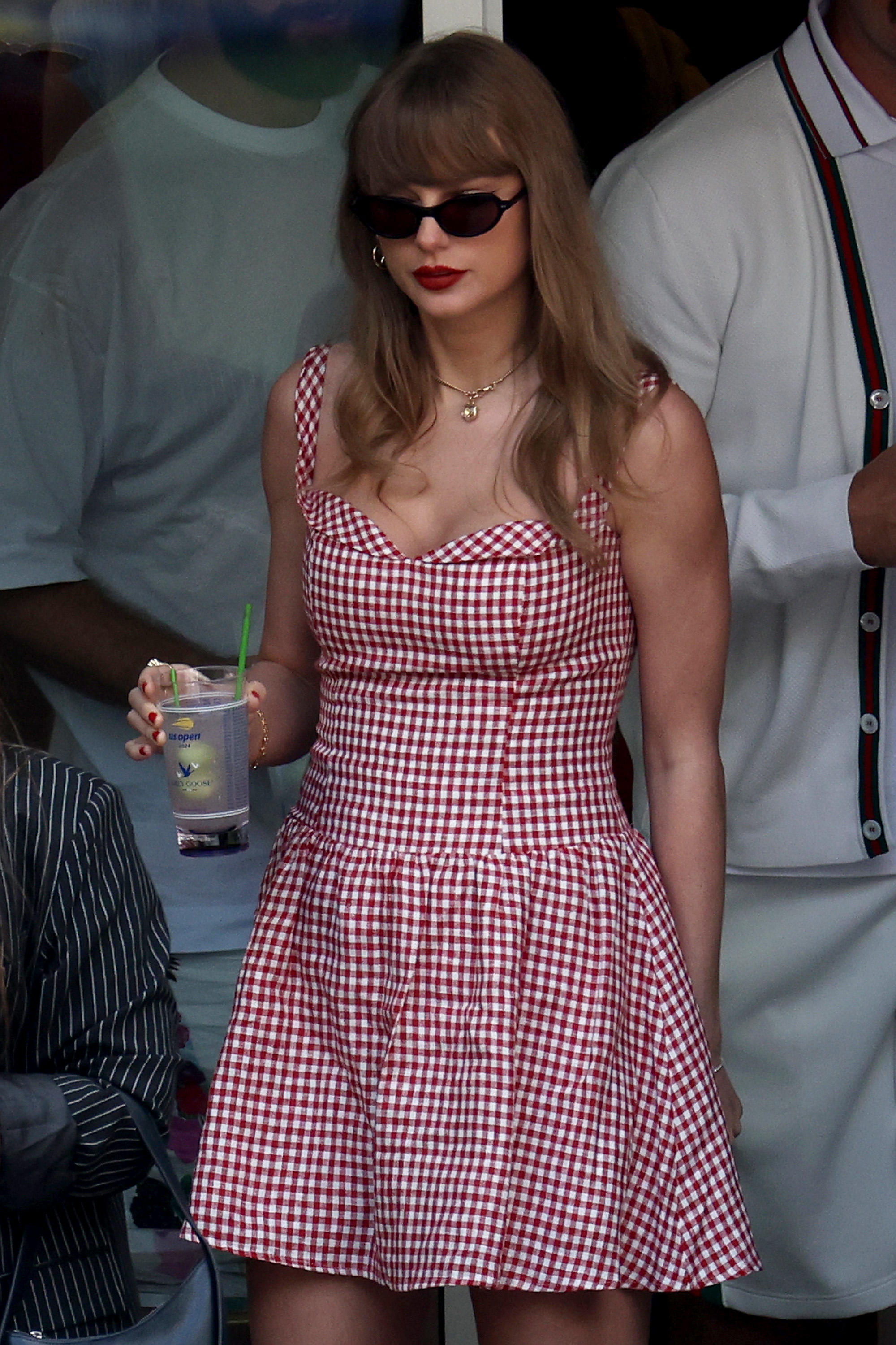 Imagem da cantora Taylor Swift usando um vestido xadrez vermelho e branco, segurando um copo de bebida.