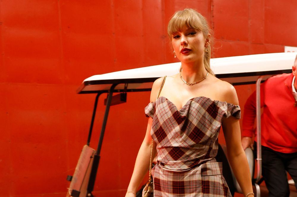 Imagem da cantora Taylor Swift usando um vestido com ombro caído xadrez em um fundo vermelho.