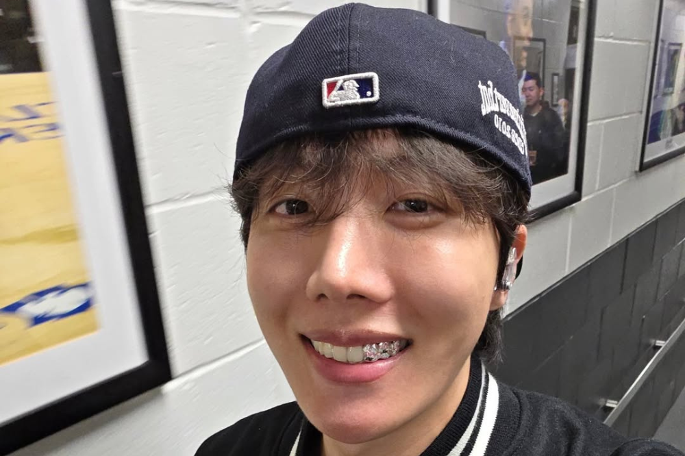 J-Hope num corredor com uns quadros no fundo. O artista está de boné e está sorrindo.